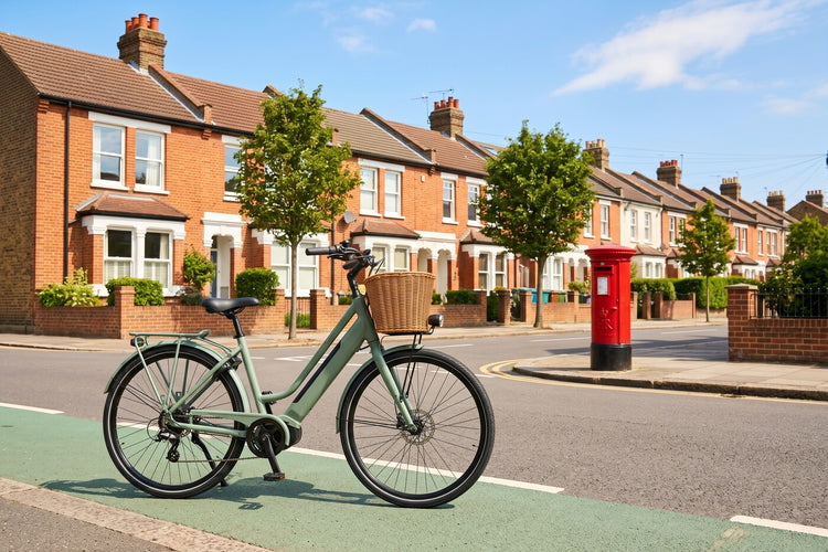 Sage-green step-through electric bike parked on a green cycle lane by British red-brick terraced houses on a sunny summer day